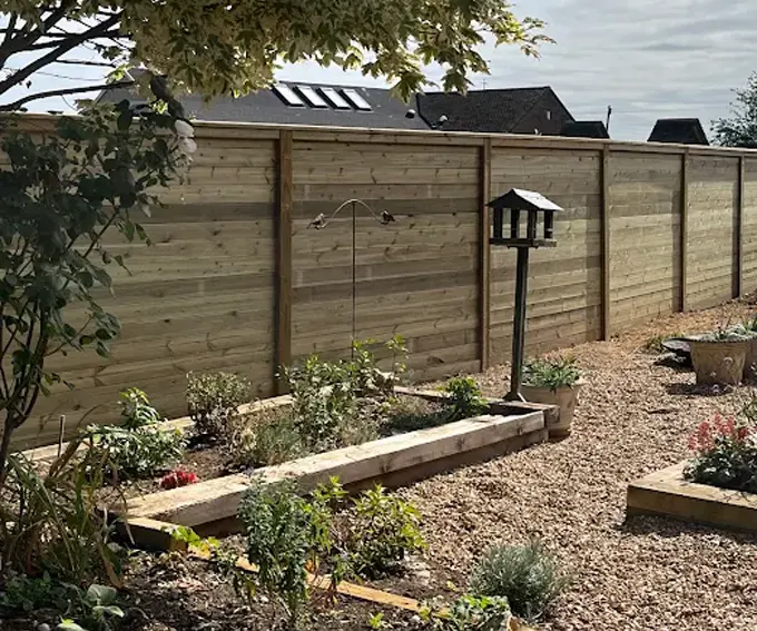 Closeboard fencing installation in a garden setting, featuring timber panels, gravel surface, and decorative bird feeder, surrounded by flowering plants and shrubs.